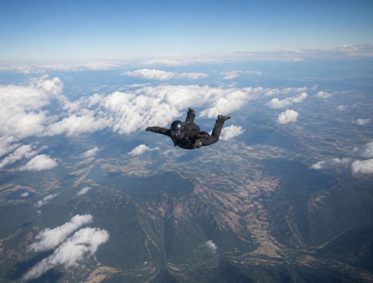Hailey Rubin skydiving in freefall above clouds and open countryside, helmet and goggles on, face not visible